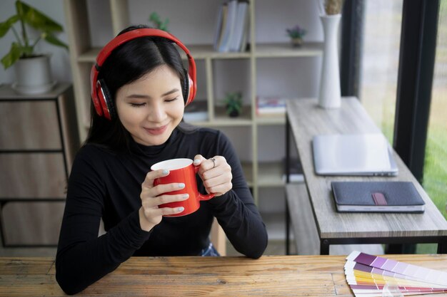Lächelnde kreative Frau, die Kopfhörer trägt und rote Kaffeetasse hält