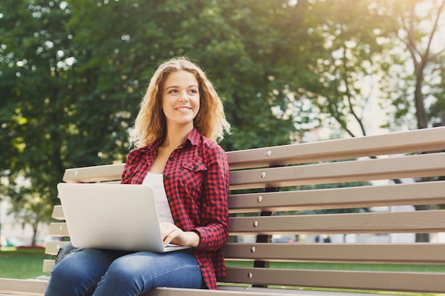 Lächelnde lässige Frau, Freiberuflerin, die mit Laptop im Park in angenehmer Atmosphäre arbeitet. Technologie-, Kommunikations-, Bildungs- und Fernarbeitskonzept, Kopierraum