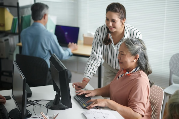 Lächelnde Lehrerin hilft einer älteren Frau beim Versenden von E-Mails und beim Installieren einer Anwendung auf dem Computer