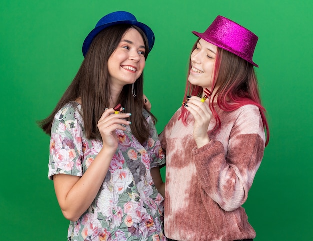 Lächelnde Mädchen mit Partyhut und Partypfeife schauen sich einzeln auf grüner Wand an