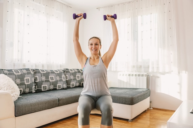 Lächelnde muskulöse Sportlerin, die auf dem Stuhl in ihrer Wohnung sitzt und Fitnessübungen für Bizeps mit Hanteln macht.