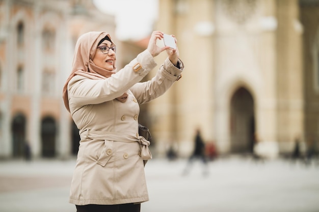 Lächelnde muslimische Frau, die einen Hijab trägt und Fotos mit ihrem Smartphone macht, während sie in der städtischen Umgebung spazieren geht.