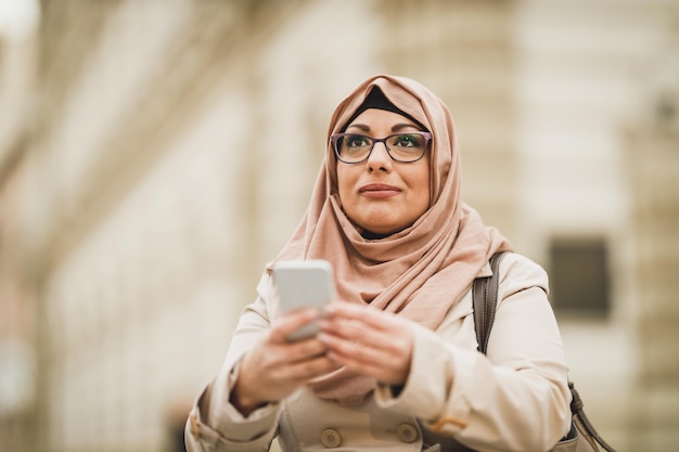 Lächelnde muslimische touristenfrau, die einen hijab trägt und ihr smartphone benutzt, während sie in einer städtischen umgebung steht.