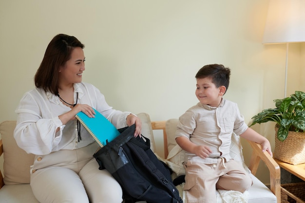 Lächelnde Mutter, die dem kleinen Sohn hilft, den Rucksack für den ersten Schultag zu packen