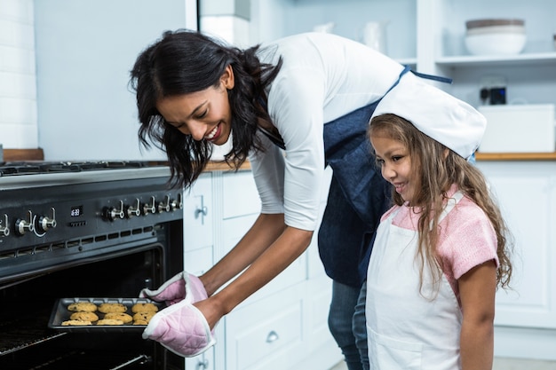 Lächelnde Mutter, die Kekse in den Ofen mit Tochter setzt