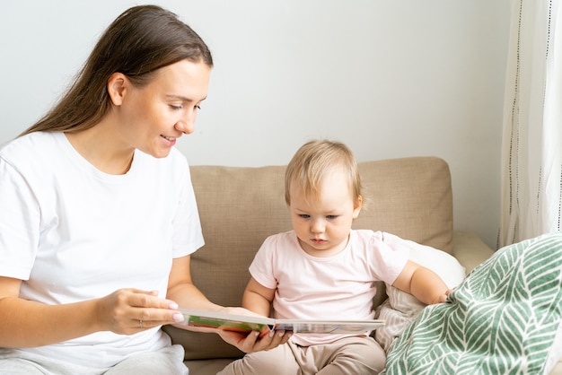 Lächelnde Mutter liest Buch mit neugierigem Kleinkind auf der Couch im Wohnzimmer mit horizontalem Tageslicht