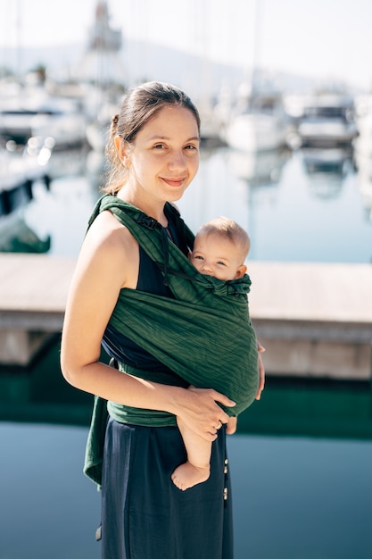 Lächelnde Mutter mit einem Baby im Tragetuch auf dem Hintergrund der Yachten closeup