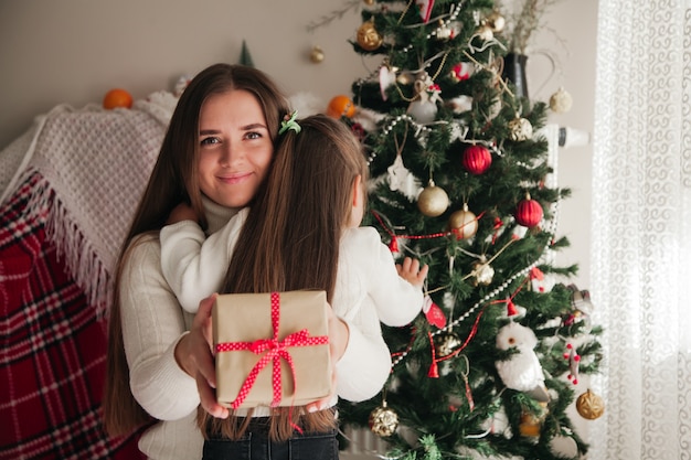 Lächelnde Mutter umarmt ihre Tochter mit Geschenken in den Händen und Weihnachtsschmuck im Hintergrund Weihnachtsfamilie präsentiert Konzept family