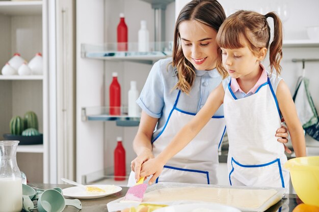 Lächelnde Mutter zeigt Tochter, wie man Silikonspatel zum Glätten von flüssigem Teig im Backblech verwendet