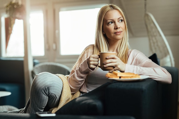 Lächelnde, nachdenkliche Frau, die sich auf dem Sofa entspannt und eine Tasse Kaffee im Wohnzimmer genießt