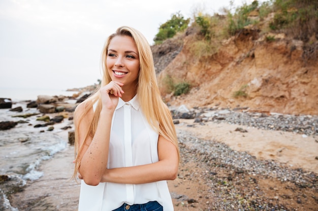 Lächelnde nachdenkliche junge Frau, die am Strand steht und denkt