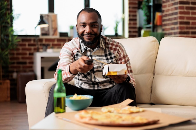 Lächelnde Person, die mit der Fernbedienung des Fernsehers die Kanäle wechselt und Nudeln von der Essenslieferung zum Mitnehmen auf der Couch isst. Spaß mit Film oder Film haben, mit Stäbchen Essen zum Mitnehmen essen.