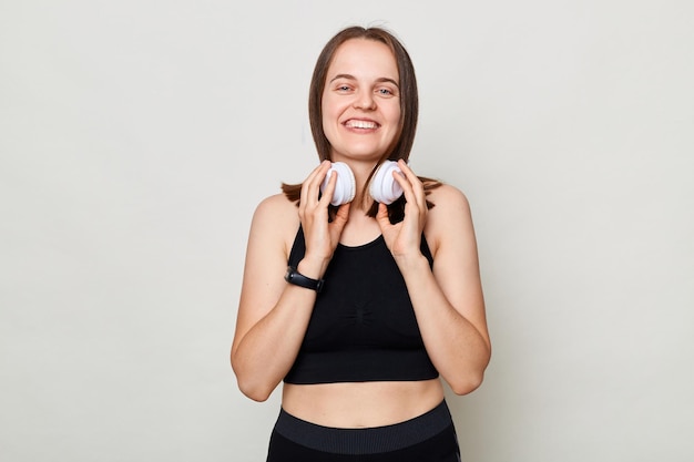 Lächelnde, positiv fröhliche, schlanke Frau mit Kopfhörern in Sportbekleidung posiert vor grauem Hintergrund und blickt mit positivem Gesichtsausdruck in die Kamera