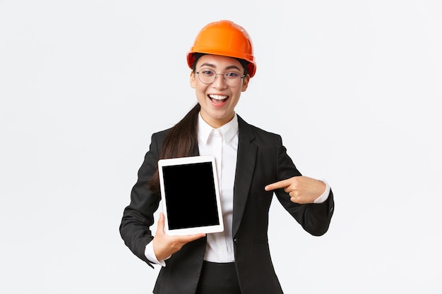 Lächelnde professionelle Bauleiterin, asiatische Ingenieurin im Schutzhelm- und Business-Anzug-Show-Projekt, die mit dem Finger auf den digitalen Tablet-Bildschirm mit zufriedenem Ausdruck zeigt, weißer Hintergrund