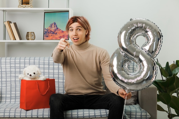 Lächelnde Punkte an der Vorderseite des hübschen Mannes am glücklichen Frauentag, der den Ballon Nummer acht hält, der auf dem Sofa im Wohnzimmer sitzt