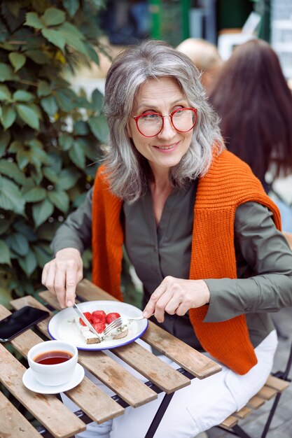 Lächelnde reife Dame isst Toast mit Sahne und Erdbeeren, die am Tisch auf der Terrasse des Cafés im Freien sitzen?