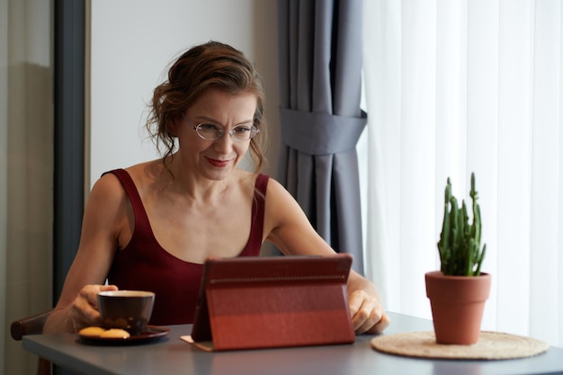 Lächelnde, reife, elegante Frau mit Brille, die Morgenkaffee trinkt und Nachrichten auf dem Tablet-Computer ansieht