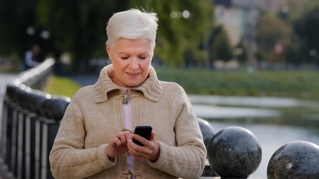 Lächelnde reife Frau aus den 60ern, die SMS-Messaging auf modernem Smartphone-Gadget sendet, glückliche Seniorin der 70er Jahre, die sich im Freien ausruht, surfen Sie im Internet auf dem Handy, überprüft E-Mails, tägliches, einfaches Nutzungstechnologiekonzept