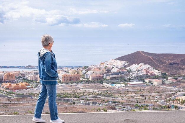 Lächelnde reife Frau Denim gekleidet stehend auf dem Bürgersteig am Handy sprechen. Stadt und Meereslandschaft im Hintergrund