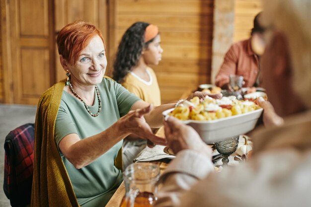 Lächelnde reife Frau, die hausgemachtes Essen einnimmt, das von ihrem Ehemann gehalten wird, während sie hilft, einen festlichen Tisch für ein Familienessen zu servieren?
