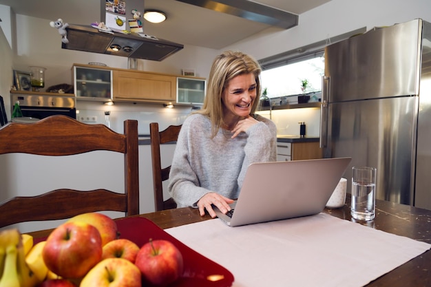 Lächelnde reife Frau mit Laptop, während sie zu Hause sitzt