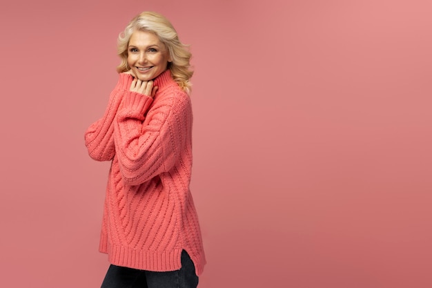 Lächelnde reife Frau mit warmem Winterpullover isoliert auf rosa Hintergrund