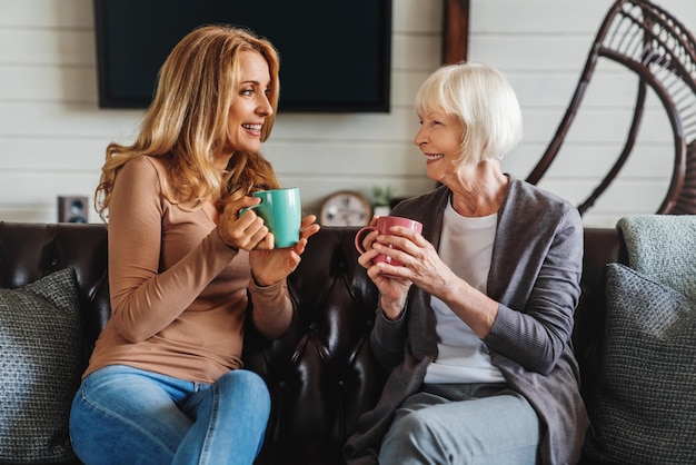 Lächelnde reife Mutter und erwachsene Tochter auf dem Sofa im Wohnzimmer sprechen und trinken Tee oder Kaffee