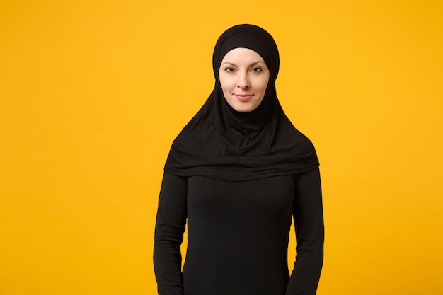 Lächelnde ruhige junge arabische muslimische Frau im Hijab bedeckt ihr Haar schwarze Kleidung isoliert auf gelber Wand, Porträt. Menschen religiöses Lifestyle-Konzept. Mock-up-Kopierraum