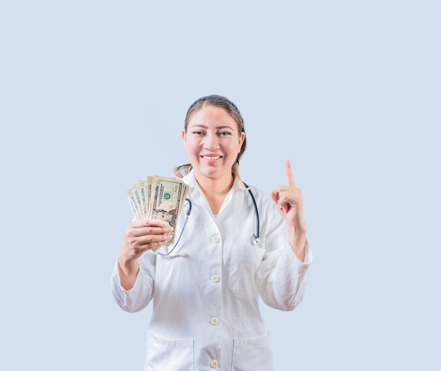Lächelnde Ärztin hält Geld in der Hand und zeigt isoliert nach oben Fröhliche junge Ärztin zeigt Geld und zeigt auf Promo nach oben