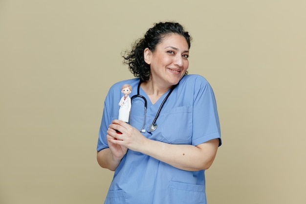 Lächelnde Ärztin mittleren Alters in Uniform und Stethoskop um den Hals, die eine Arztfigur mit beiden Händen hält, die isoliert auf olivgrünem Hintergrund in die Kamera schauen