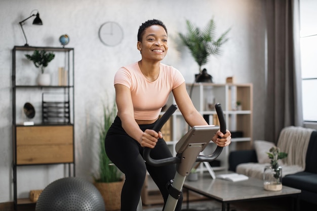 Lächelnde schöne afrikanisch-amerikanische Sportlerin in Sportbekleidung mit Fahrrad zu Hause