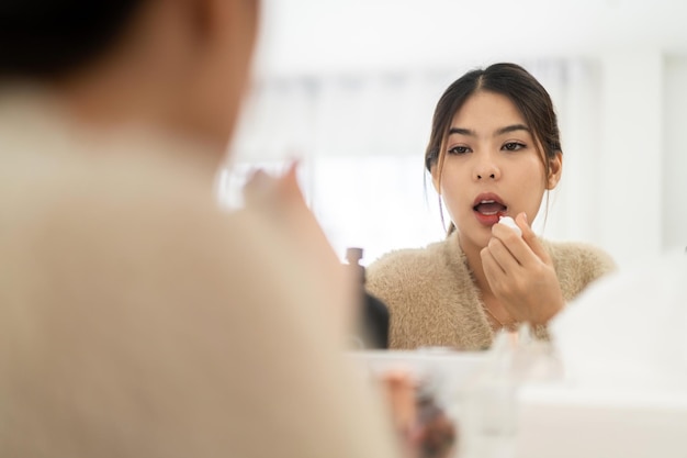 Foto lächelnde schöne asiatische frau frische gesunde haut, die auf spiegel schaut und hautpflegende lippen mit rotem lippenstift auf homefacial schönheits- und kosmetikkonzept aufträgt