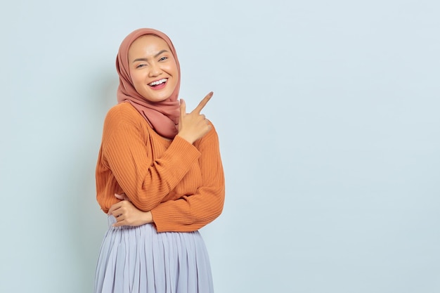 Lächelnde schöne asiatische Frau in braunem Pullover und Hijab, die mit dem Finger in den Kopierraum zeigt, der Werbeprodukte zeigt und selbstbewusst in die Kamera blickt, isoliert über weißem Hintergrund