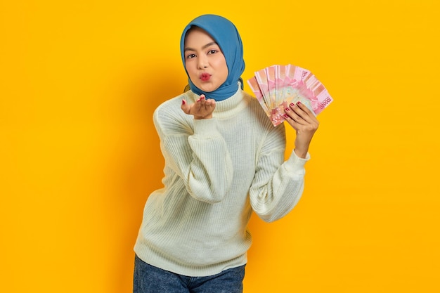 Lächelnde schöne asiatische Muslimin in weißem Pullover, die Bargeld in indonesischen Rupiah-Banknoten hält und Handflächen hält, senden einen Luftkuss über gelben Hintergrund ​Menschen religiöses Lebensstilkonzept