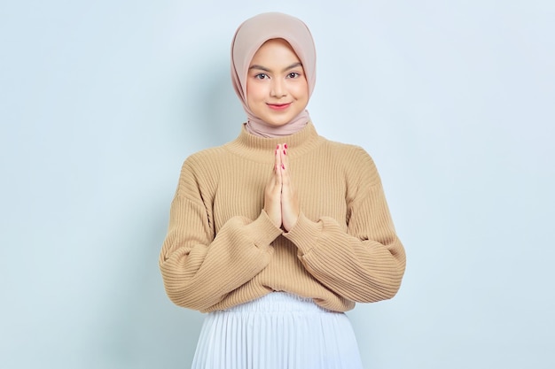 Lächelnde schöne asiatische muslimische Frau im braunen Pullover, die Eid Mubarak-Gruß gestikuliert, isoliert über weißem Hintergrund Religiöses Lifestyle-Konzept der Menschen