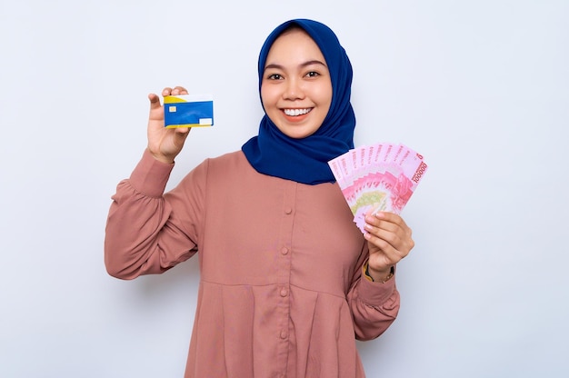 Lächelnde schöne asiatische muslimische Frau in einem lässigen Hemd und Hijab mit indonesischen Rupiah-Banknoten, die eine Kreditkarte zeigen, die über weißem Hintergrund isoliert ist Religiöses Lifestyle-Konzept der Menschen