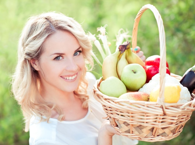Lächelnde schöne blonde Frau, die Strohkorb mit frischen Früchten im Freien über Naturhintergrund hält