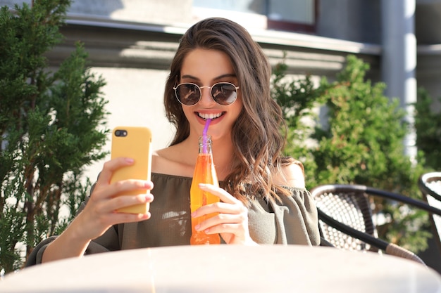 Lächelnde schöne brünette Frau, die im Straßensommercafé sitzt, Selfie mit dem Handy machen und Saft trinken.