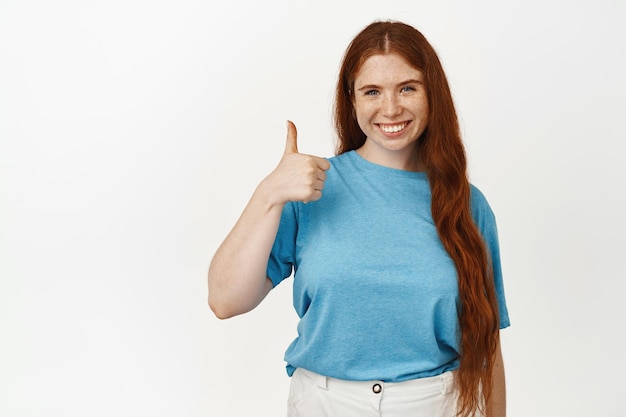 Lächelnde schöne Frau mit roten Haaren, Ingwermädchen zeigt Daumen nach oben zur Zustimmung, Unterstützung und Zustimmung, Lob für gute Wahl, Kompliment, gut gemacht, Geste für gute Arbeit, vor weißem Hintergrund stehend