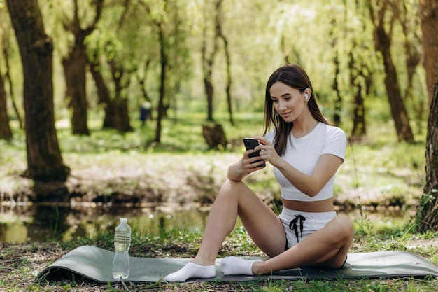 Lächelnde schöne Frau mit Telefon beim Sport am Morgen in der Parknatur auf der Matte mit rosafarbenem Hemd, Fitness-Outfit, glücklicher, gesunder Lebensstil, Musik im Ohrhörerfahrrad im Hintergrund