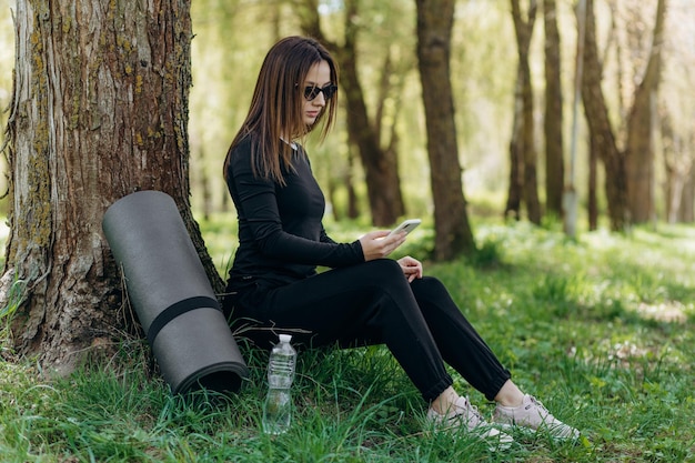 Lächelnde schöne Frau mit Telefon beim Sport am Morgen in der Parknatur auf der Matte mit rosafarbenem Hemd, Fitness-Outfit, glücklicher, gesunder Lebensstil, Musik im Ohrhörerfahrrad im Hintergrund