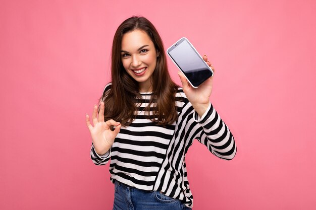 Lächelnde schöne glückliche junge Frau mit gestreiftem Pullover isoliert über dem Hintergrund mit Kopienraum, der eine ok Geste zeigt, die auf die Kamera blickt, die die Bildschirmanzeige des Mobiltelefons zeigt. Mock-up, Ausschnitt