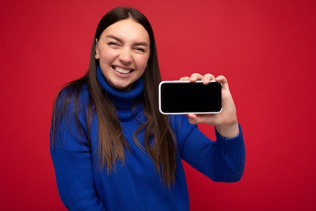 Lächelnde schöne glückliche junge Wandfrau, die lässigen blauen Pullover lokalisiert über Rot trägt