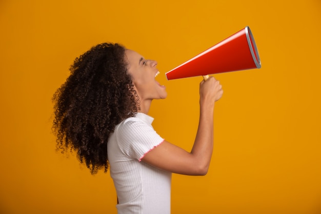 Lächelnde schöne junge Afroamerikanerin mit lockigem Afro-Haar, das durch Megaphon über gelber Wand schreit