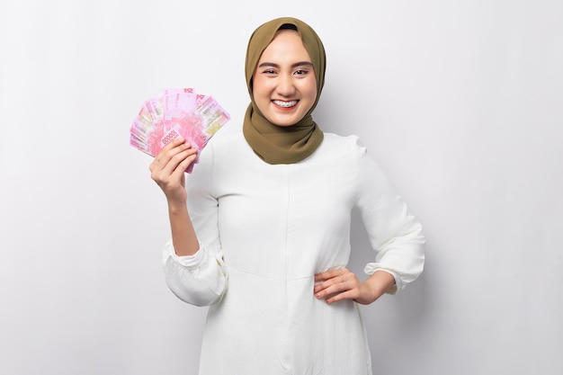 Lächelnde schöne junge asiatische muslimische Frau mit Hijab, die Bargeld in Rupiah-Banknoten mit der Hand hält, während sie die Kamera isoliert auf weißem Hintergrund betrachtet Religiöses Lifestyle-Konzept der Menschen