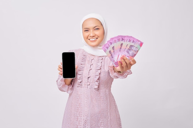 Lächelnde schöne junge asiatische muslimische Frau mit Hijab und lila Kleid, die ein Handy mit leerem Bildschirm zeigt und Bargeld-Rupiah-Banknoten isoliert auf weißem Studiohintergrund hält