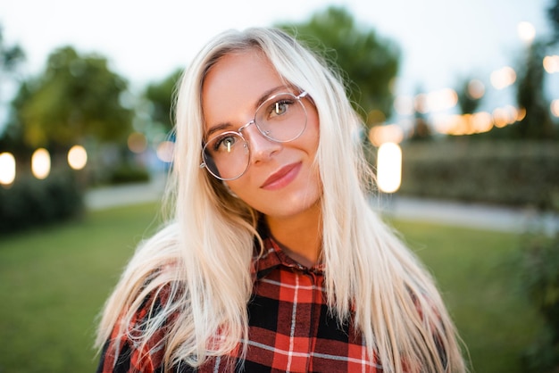 Lächelnde schöne junge erwachsene Studentin tragen eine Brille über leuchtenden Lichtern und naturgrünem Hintergrund im Freien in der Nähe Blick in die Kamera Zurück zur Schule