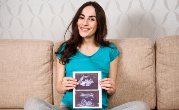 Lächelnde schöne schwangere Frau, die auf der Couch sitzt und ein Ultraschallbild hält und es der Kamera zeigt Warten auf das Kind