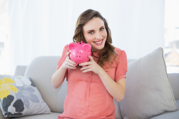 Lächelnde schwangere Frau, die das rosa Sparschwein sitzt auf Couch rüttelt