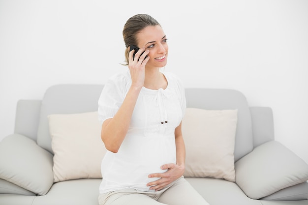 Lächelnde schwangere Frau, die mit ihrem Smartphone sitzt auf Couch anruft